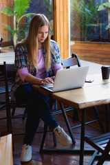 Blonde girl sitting in cozy hipster cafe with laptop and coffee cup. Woman freelancer typing on a netbook indoors.