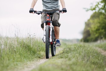 Low angle view of young man riding bicycle on a meadow path with green grass. Environmentally friendly transport, clean air, activity, healthy lifestyle concept