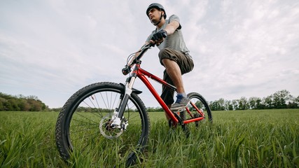 Fototapeta na wymiar Bicycle sports, traveling, healthy lifestyle and activity. Low angle view of young man riding bicycle on a meadow with high green grass