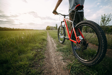 Fototapeta premium Low angle view of young man in helmet standing with bicycle at country road enjoying sunset. Tourism, healthy lifestyle and activity