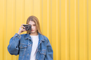 Attractive hipist girl photographed on a film camera on a yellow background. Girl in a jeans jacket on a yellow background with a film camera