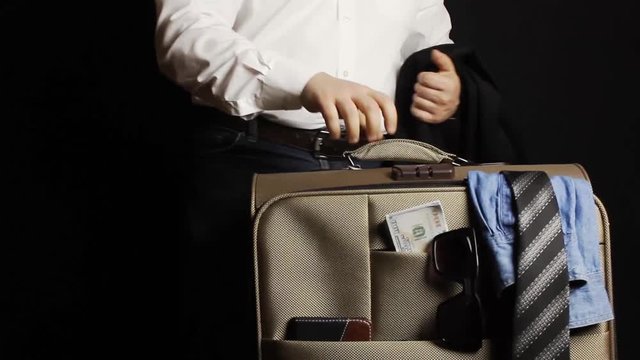 Businessman in jeans and shirt bringing in a travel suitcase with money, glasses & clothing and standing with it on black background.