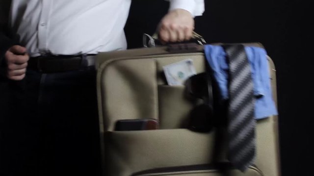 Businessman in jeans and shirt taking away a travel suitcase with money, glasses & clothing on black background.