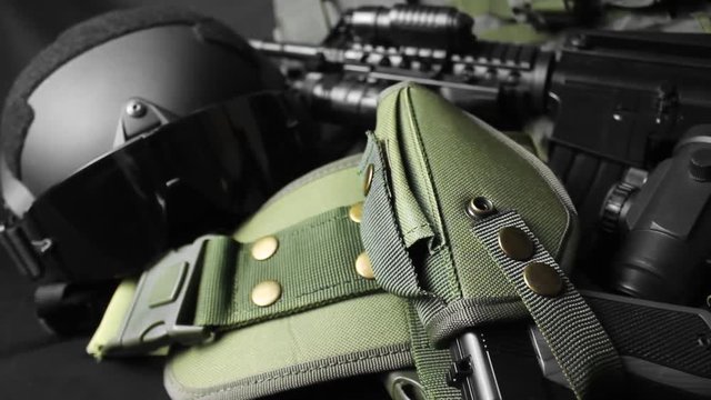 Photo of a tactical military vest, rifle, gun, helmet with glasses close-up view and cartrige belt laying on black cloth table background.