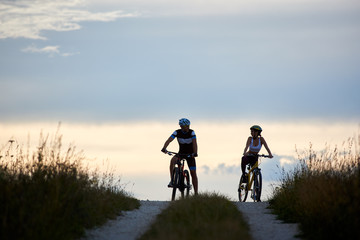 Obraz premium Couple of cyclists sitting on bicycles on a country road watching each other in the background a horizon with a beautiful evening sky. The guys are dressed in sports clothes and helmets