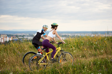Fototapeta na wymiar Two cyclists ride on a hill in the grass with wild flowers in the distance the city and mountains are moving. The man looks at the woman with a smile. Both are dressed in sports clothes and helmets