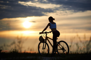 Fototapeta na wymiar Back view of slender female cyclists sitting on bicycle and posing in sportswear. Sporty woman riding bike, enjoying and observing amazing and fantastic view of sunset and landscape.