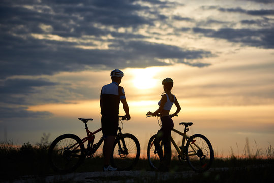 Back view of a couple of cyclists standing with bicycles and enjoying the sunset. A guy and a girl are dressed in helmets and sportswear. Perfect sky with clouds and evening sun