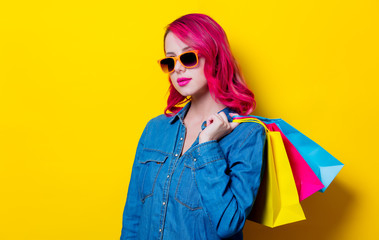 Young pink hair girl in sunglasses and blue shirt holding a colored shopping bags. Portrait on isolated yellow background