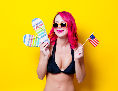 Young pink hair girl in bikini and orange glasses with flip flops and american flag. Portrait isolated on yellow background