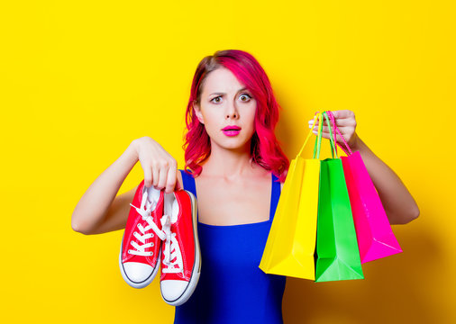 Young pink hair girl in blue dress with colored shopping bags and gumshoes. Portrait isolated on yellow background