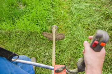Man wearing overalls and rubber boots mowing green grass with gasoline lawn trimmer. The...