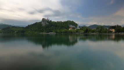 Panoramiczny widok Bled, Słowenia, jezioro, skaliste wzgórze z ruinami  średniowiecznego zamku, poniżej wieże kościoła, zieleń i niebo odbija się w spokojnej tafli wody - obrazy, fototapety, plakaty