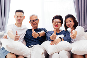 Asian family with adult children and senior parents giving thumbs up and relaxing on a sofa at home...