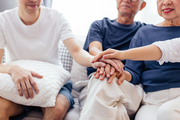 Close up of family with adult children and senior parents putting hands together and sitting on sofa at home together. Family unity and cooperation concept
