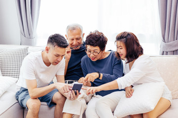 Asian family with adult children and senior parents using a mobile phone and relaxing on a sofa at home together