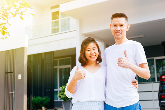 Asian Couple Standing In Front Of Their New House And Giving Thumbs Up. Newlyweds Family Just Moved In New Home And Car Carrying Boxes