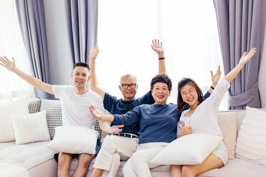 Asian Family With Adult Children And Senior Parents Raising Hands Up And Sitting On A Sofa At Home. Happy And Relaxing Family Time