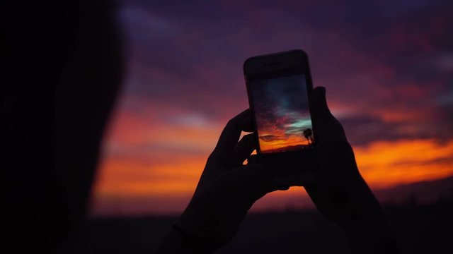 Silhouette of a young girl taking a selfie and shooting pictures with her smart phone of an orange sunset with dramatic colors in Santa Monica Beach, California.