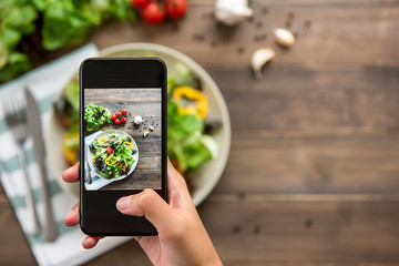 Hand holding smartphone taking photo of beautiful food, mix fresh green salad on wood table, to...