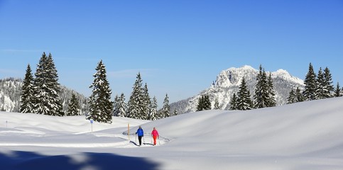 Two hikers walk through snow-covered mountain scenery, premium winter hiking trail, Hemmersuppenalm, Reit im Winkl, Chiemgau, Oberbayern, Bavaria, Germany.