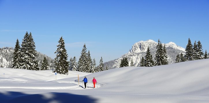 Two hikers walk through snow-covered mountain scenery, premium winter hiking trail, Hemmersuppenalm, Reit im Winkl, Chiemgau, Oberbayern, Bavaria, Germany.