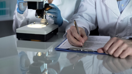 Lab worker filling paper forms, his assistant viewing samples under microscope