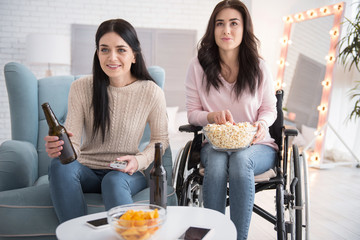 Little pause. Optimistic sister and disabled woman watching movie and eating popcorn with beverage