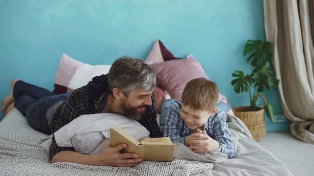 Caring father is reading funny book to his child while boy is laughing and talking to his parent. Full-size bed, bright pillows and green plants are visible.