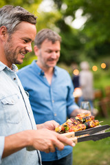 in a summer evening,  two men  in their forties prepares a barbecue for  friends gathered around a table in the garden