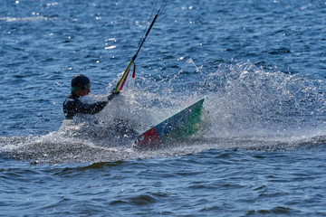Obraz premium A male kiteboarder rides on a board on a large river. He performs various exercises while moving on water. Splashes of water scatter in different directions. The sun's rays shine in the water. 