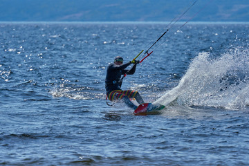 A male kiteboarder rides on a board on a large river. He performs various exercises while moving on water. Splashes of water scatter in different directions. The sun's rays shine in the water. 