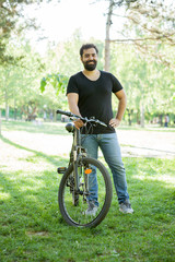 Relaxed man smiling to the camera while holding a bicycle in the park