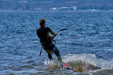 A male kiteboarder rides on a board on a large river. He performs various exercises while moving on water. Splashes of water scatter in different directions. The sun's rays shine in the water. 