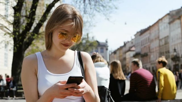outside portrait of attractive young blonde woman using smartphone looking at display and laughing internet communication typing message chatting online smiling girl sunny warm day city street