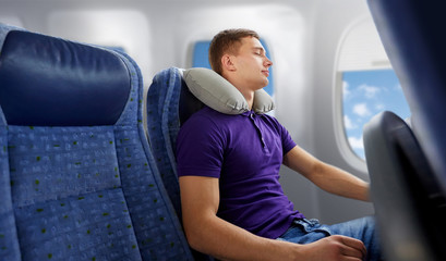 travel, tourism and people concept - happy young man sleeping in plane with inflatable pillow over...