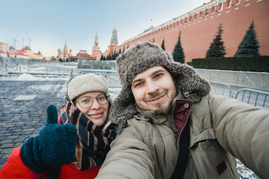 happy romantic couple of tourists in the winter in warm clothes and hats makes self-portrait selfie in front of the Kremlin on Red Square in Moscow, traveling in Russia.