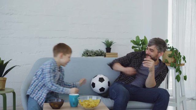 Adorable little kid is playing with ball with his active caring father, throwing and catching football on couch at home. Family bonding and funny sport concept.