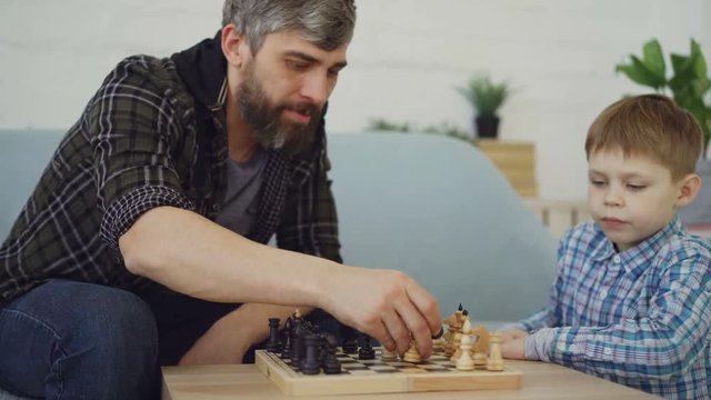 Clever small boy is playing chess with his caring father touching chesspieces and moving chessmen. Intellectual game, happy family and generations concept.