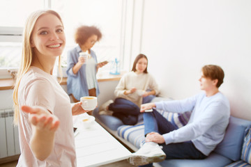 Nice picture of blonde that is looking to camera and smiling. She is pointing on it and holding a cup of coffee while her friends are talking with each other and drinking coffee.
