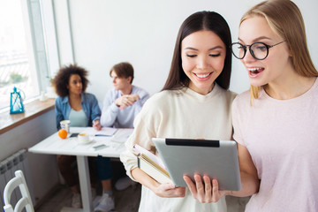 Two smart girls are standing and looking on tablet. The blonde girl wears glasses. Her friends is holding a notebook. There are their friends sitting behind them and studying.