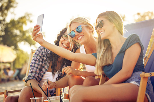 Group of young casual female friends sitting on beach on sunbeds,hangout and relaxing.Taking selfie with smart phone.