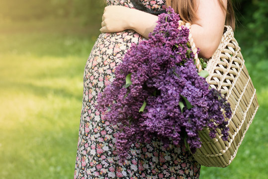 Close-up of torso of young pregnant model standing with lilac flower in park. Future mom expecting baby walking outdoors. Healthy maternity concept.