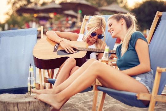 Two young female friends sitting on beach on sunbeds,singing and playing guitar.