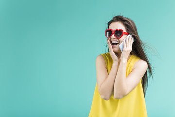 Fototapeta premium Beautiful smiling brunette girl wears yellow t-shirt and sunglasses talking by phone before blue background