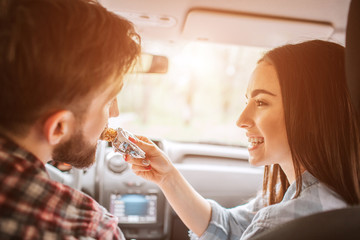 Girl is holding a sweet bar and feeding it to her boyfriend. Guy is biting a piece of bar. She is looking at him and smiling. They made a stop to eat food.