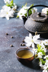 Obraz na płótnie Canvas Traditional ceramic cup of hot green tea with black iron teapot, spring flowers white magnolia and cherry blooming branches over dark blue texture background. Copy space.