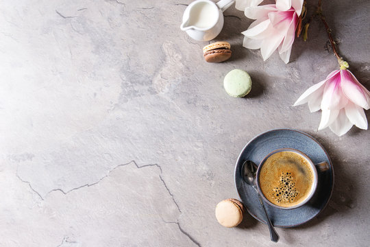 Blue cup of black espresso coffee with french dessert macaroons, cream and spring flowers magnolia branches over grey texture background. Top view, space. Spring greeting card, wallpapers