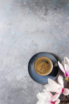 Blue cup of black espresso coffee and spring flowers magnolia branches over grey texture background. Top view, copy space. Spring greeting card