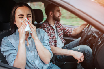 A picture of sick girl riding in car with young man. She is sneezing in napkin while he is paying attention to the road. Girl is suffering.